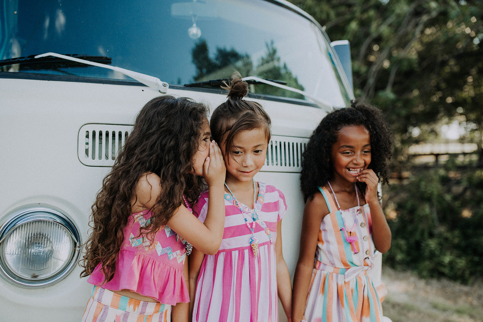 Three young girls standing in front of a vintage van, with one girl whispering to another.